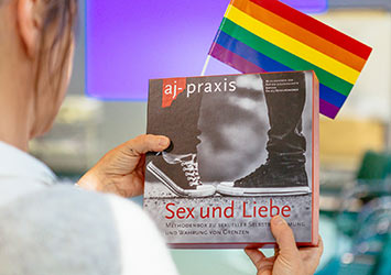 Frau hält ein Buch zum Thema Sexualität in der Hand und eine Fahne in Regenbogenfarben.